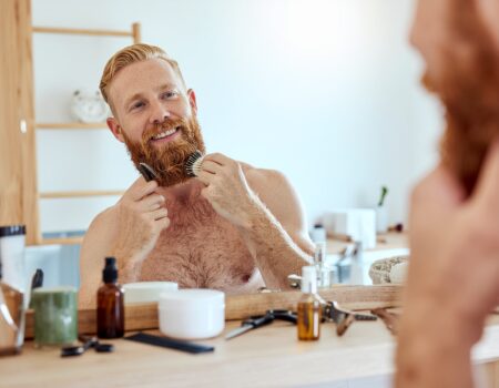 Man grooming his beard in front of a mirror, with beard care tools like a trimmer, comb, oil, and moisturizer arranged neatly on the counter