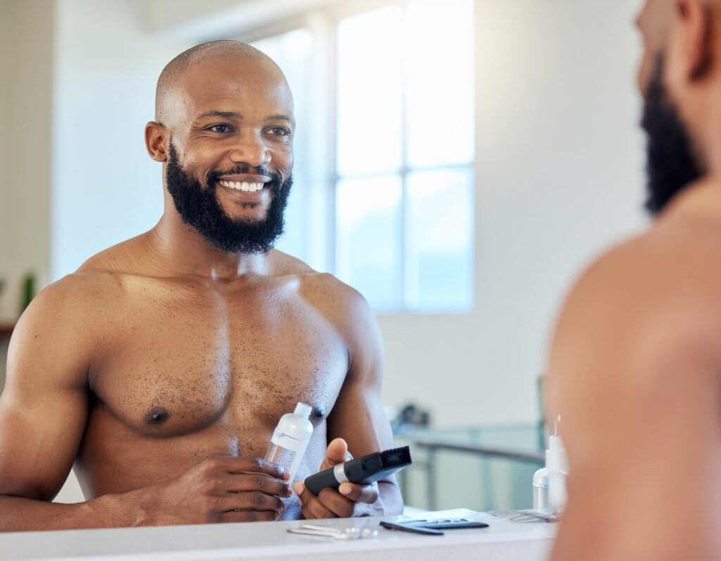 Man with a well-groomed beard standing in a modern bathroom, holding a trimmer and beard oil, surrounded by grooming tools on the counter.