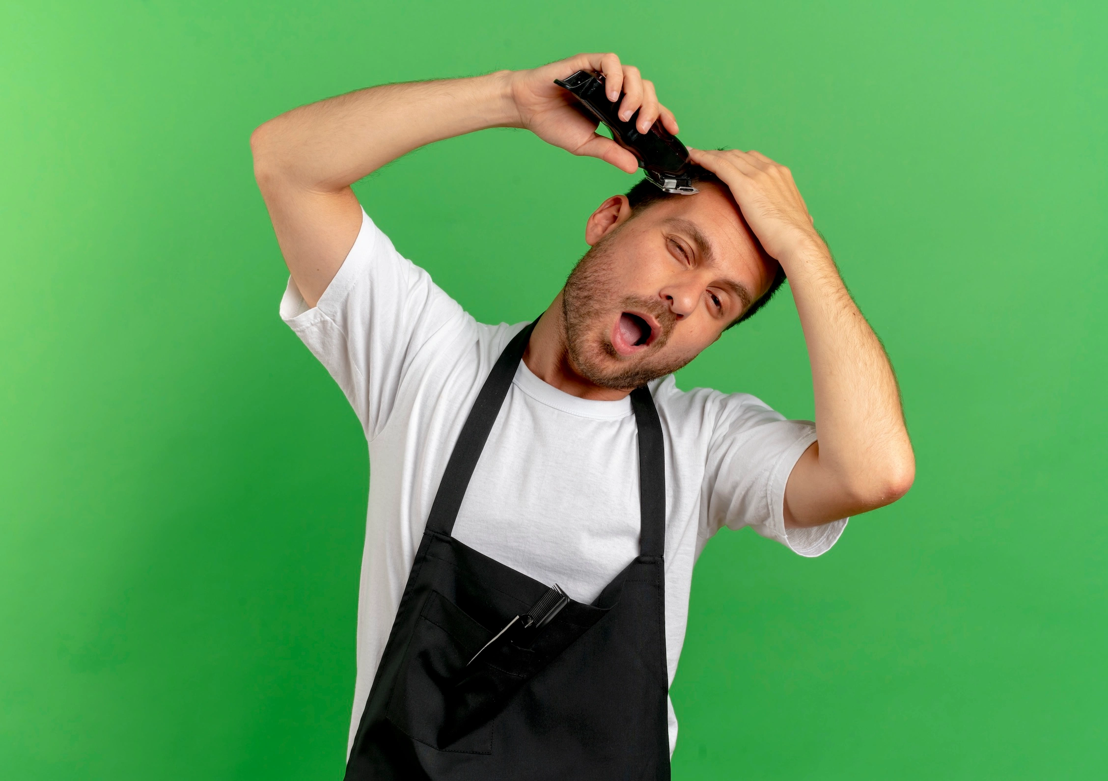 barber man apron shaving his hair with hair cutting machine looking confident standing green wall