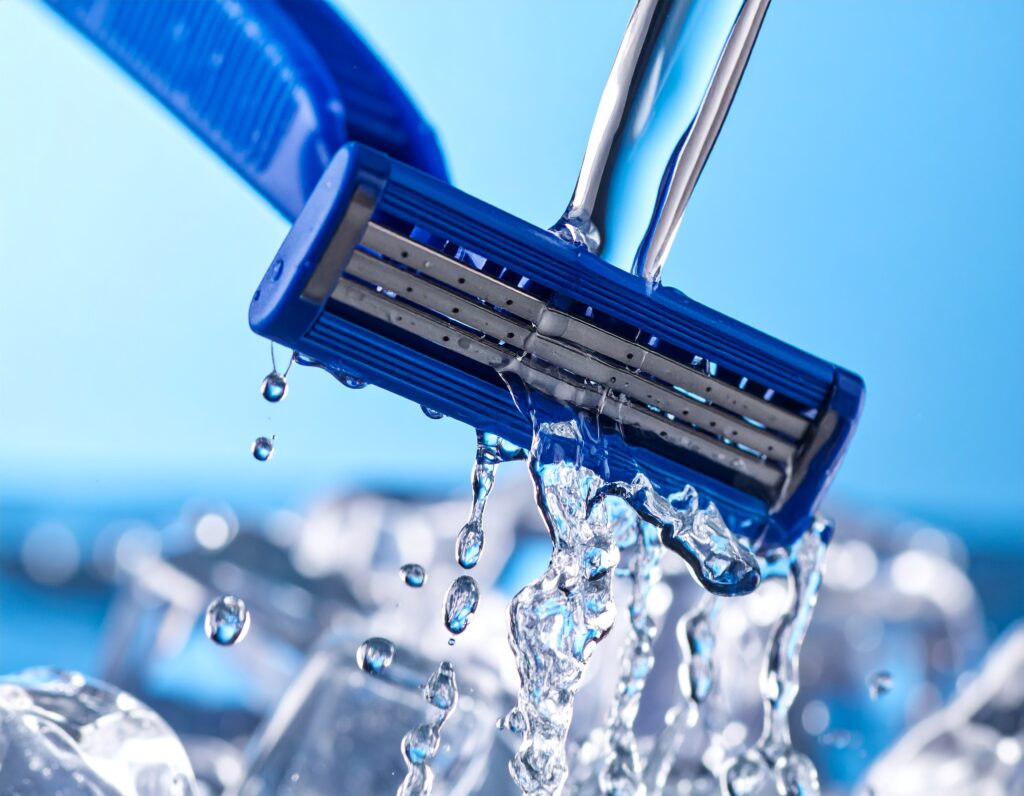 close—up of a razor being rinsed under running tap water, with clear water droplets and a shiny, clean blade.