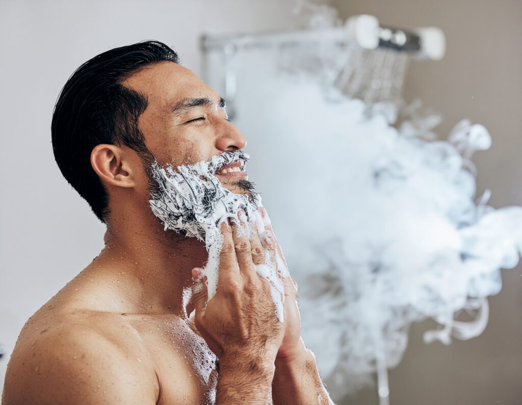 a bearded man in a modern shower, lathering beard wash into his full beard with water cascading down, steam visible in the background