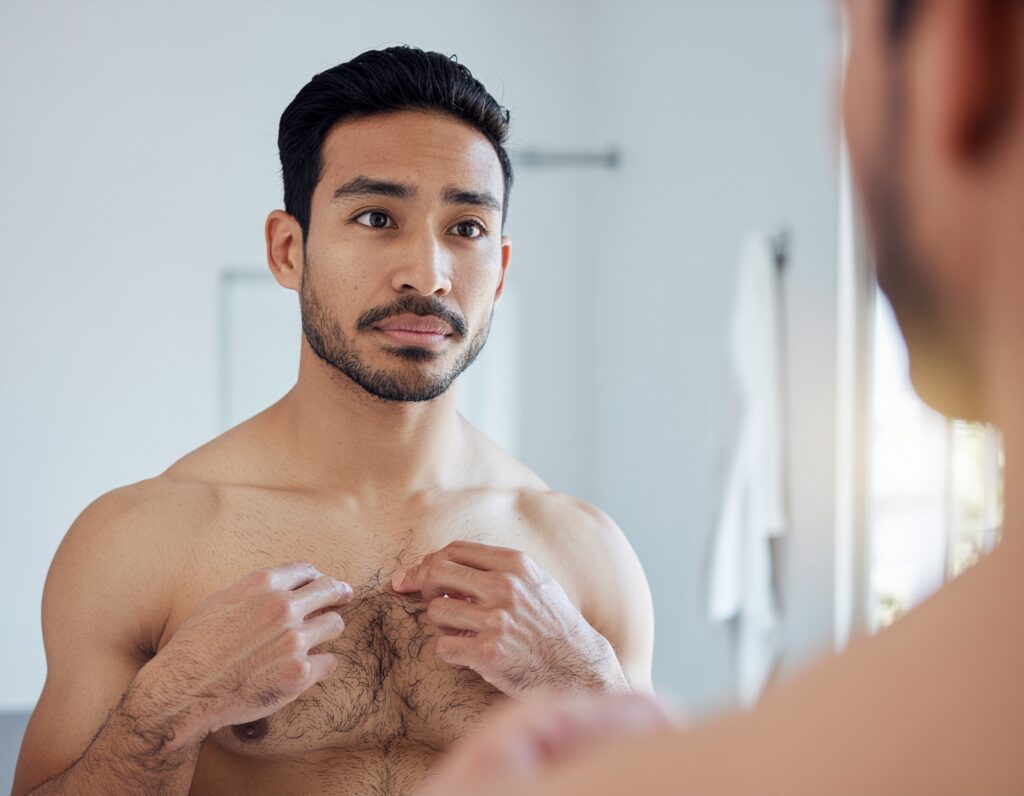Man closely inspecting his chest hair pattern in a bathroom mirror, noting areas of different growth directions and thicknesses
