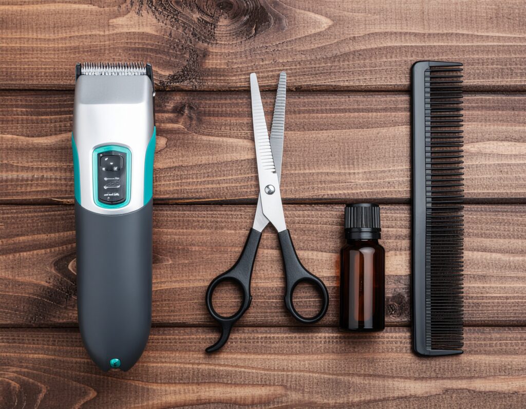 overhead view of grooming essentials arranged on wood surface: electric trimmer, precision scissors, beard oil bottle, fine—tooth comb, and natural bristle brush