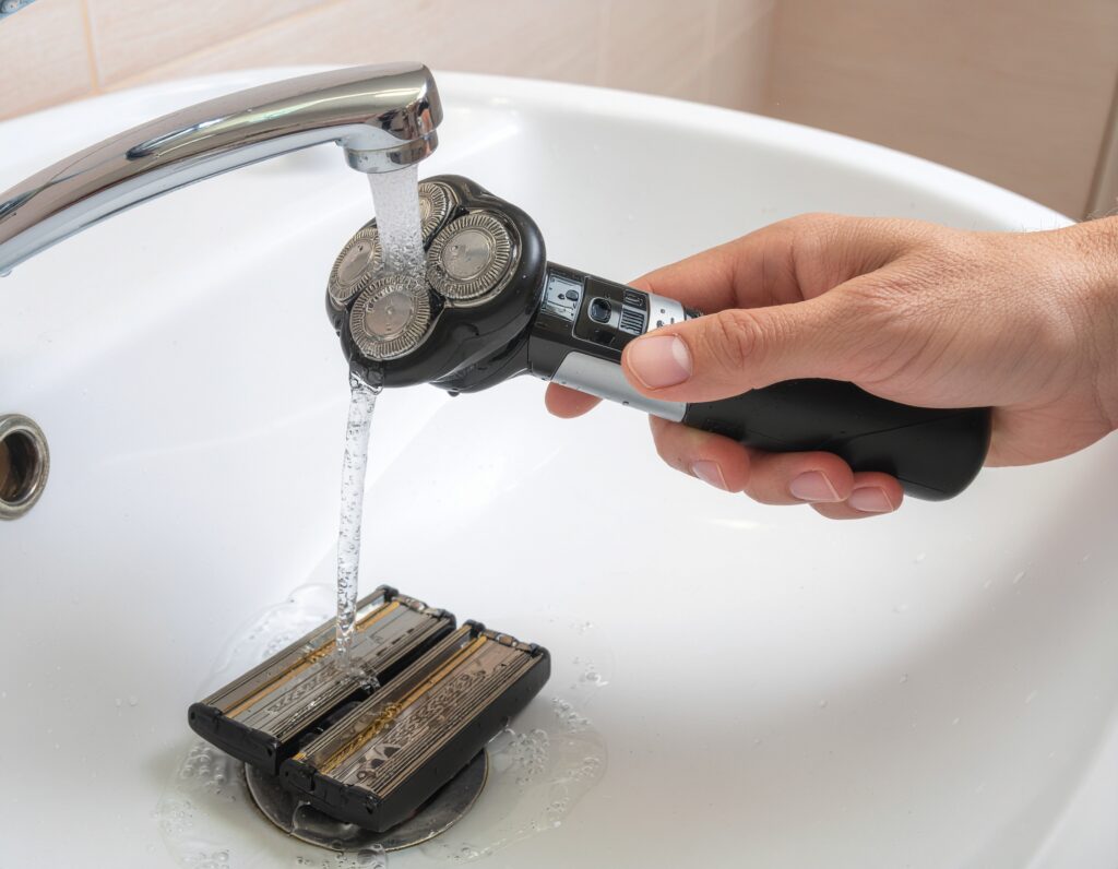 Disassembled foil shaver being rinsed under running water, with parts neatly laid out on a white bathroom counter