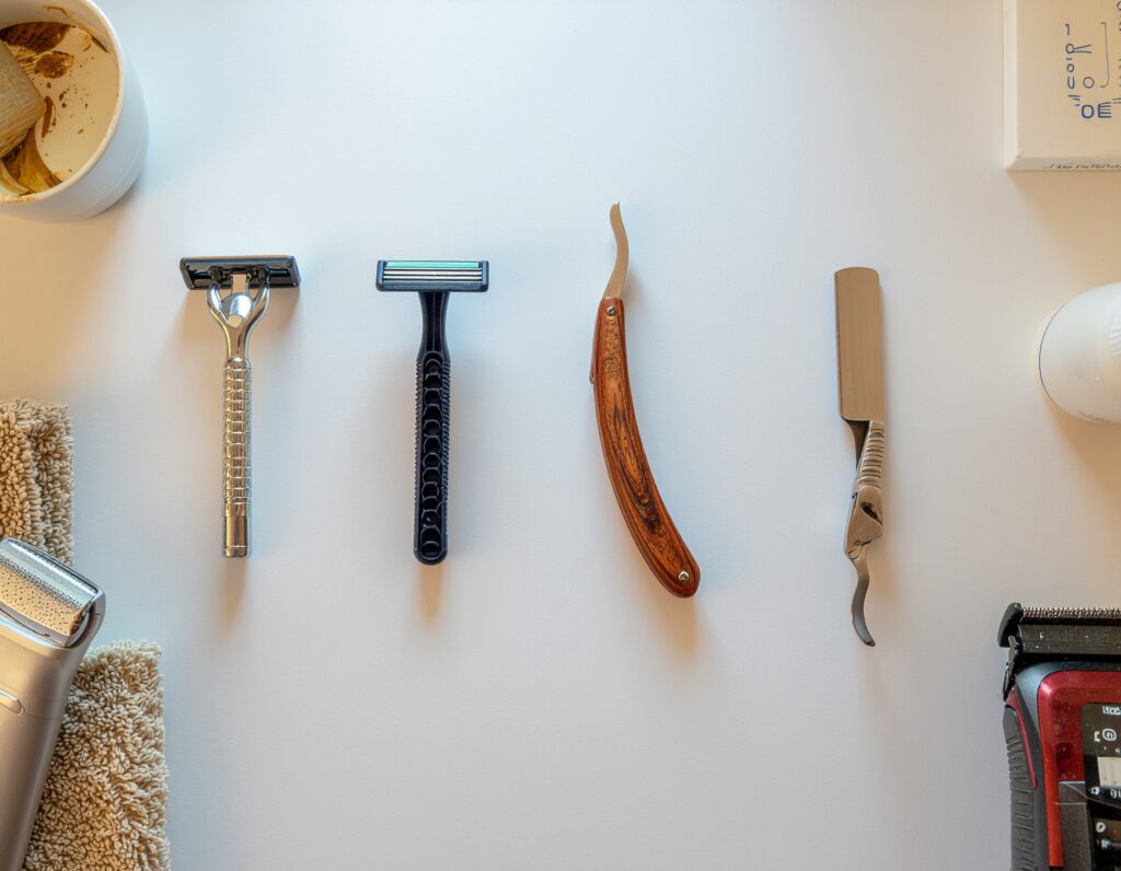 Various types of men's razors arranged on a bathroom counter — safety, cartridge, straight, and electric — alongside grooming essentials like shaving cream and a towel.