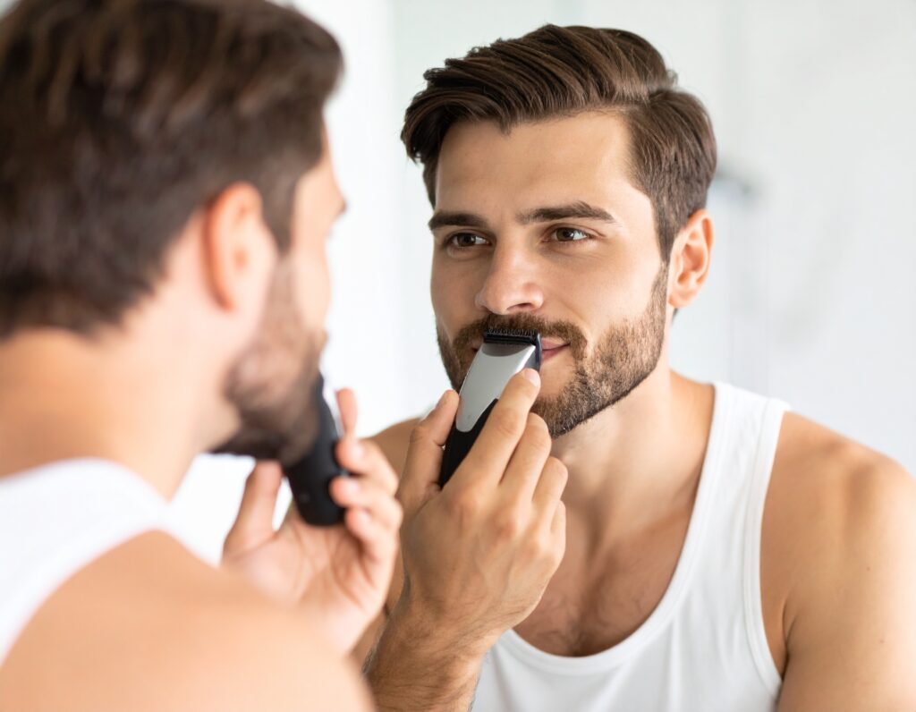 a well—groomed man in his early 30s confidently trimming his moustache with an electric trimmer while looking in a bright bathroom mirror, natural morning light illuminating his face